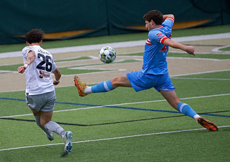 ACFC player in blue uniform with outstretched leg trying to trap ball in front of Steel City player in white uniform