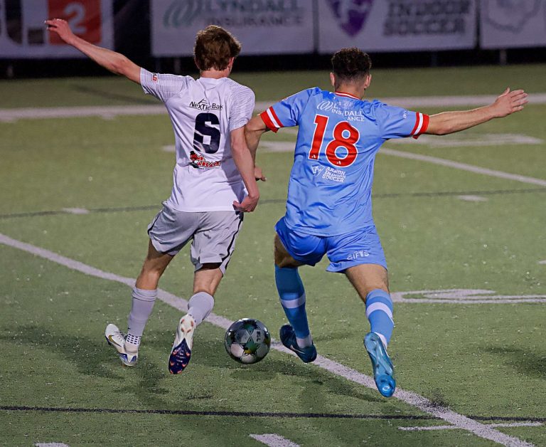 Steel City player in white uniform and ACFC player in blue uniform with outstretched arms going for the ball