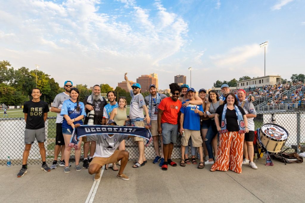 17 people in soccer supporters gear stading on concourse above soccer pitch with stands in background